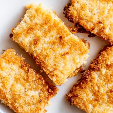 A close-up of golden brown Crispy Panko Halloumi stacked on a plate with lemon wedges and a small bowl of red hot sauce.